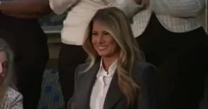 The image shows a woman in a professional setting, smiling and wearing a dark blazer over a white shirt.