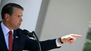 The image shows a man in a suit and tie speaking into a microphone while pointing to the side.