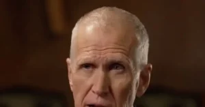 The image shows a close-up of an older man with short, light-colored hair, set against a blurred background.