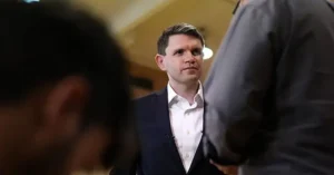 The image shows a man in a suit standing in a room, with the focus on him and the blurred figures of others around him.