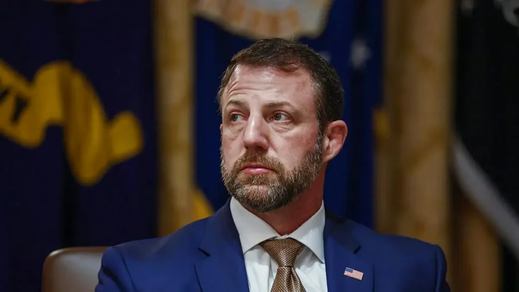The image shows a man in a suit and tie, seated in front of a backdrop featuring a flag and a seal, suggesting a formal or official setting.