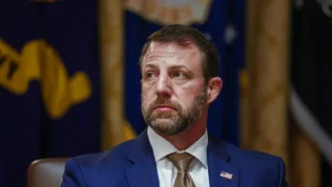 The image shows a man in a suit and tie, seated in front of a backdrop featuring a flag and a seal, suggesting a formal or official setting.