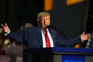 The image shows a person standing at a podium, gesturing with their hands while speaking into a microphone.