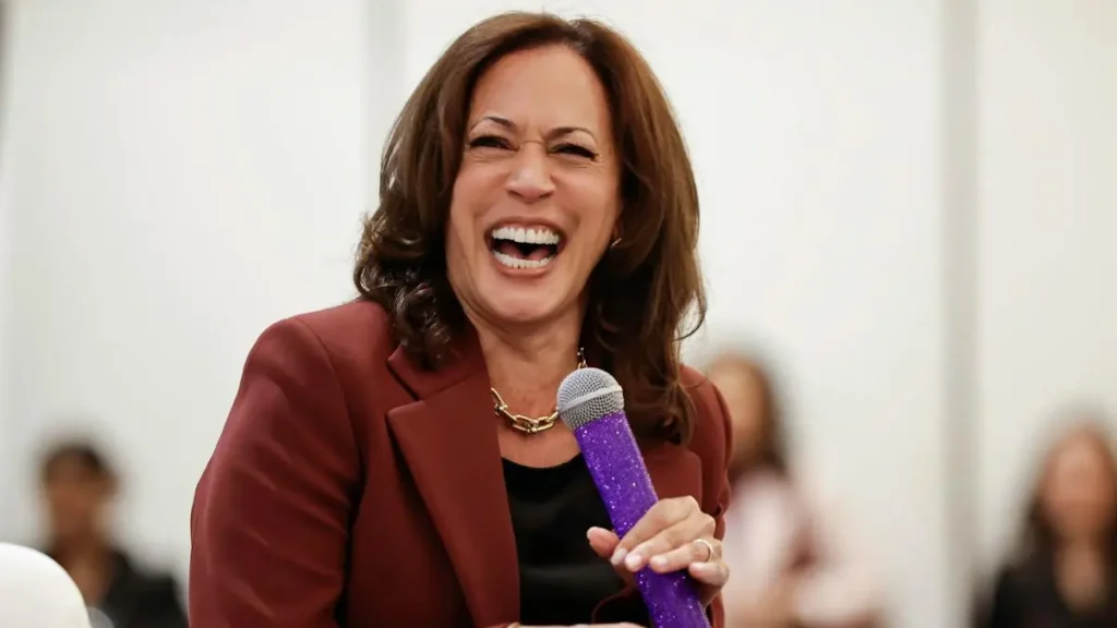 A woman in a maroon blazer is smiling and speaking into a purple microphone.