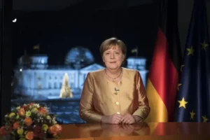 The image shows a person seated at a desk with a backdrop featuring the Reichstag building and German and European Union flags, suggesting a formal or official setting.
