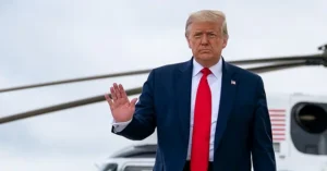The image shows a person in a suit and red tie waving, with a helicopter in the background.