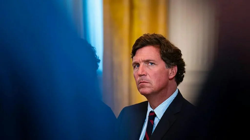 The image shows a man in a suit and tie, appearing to be in a formal setting, possibly a news conference or official event.