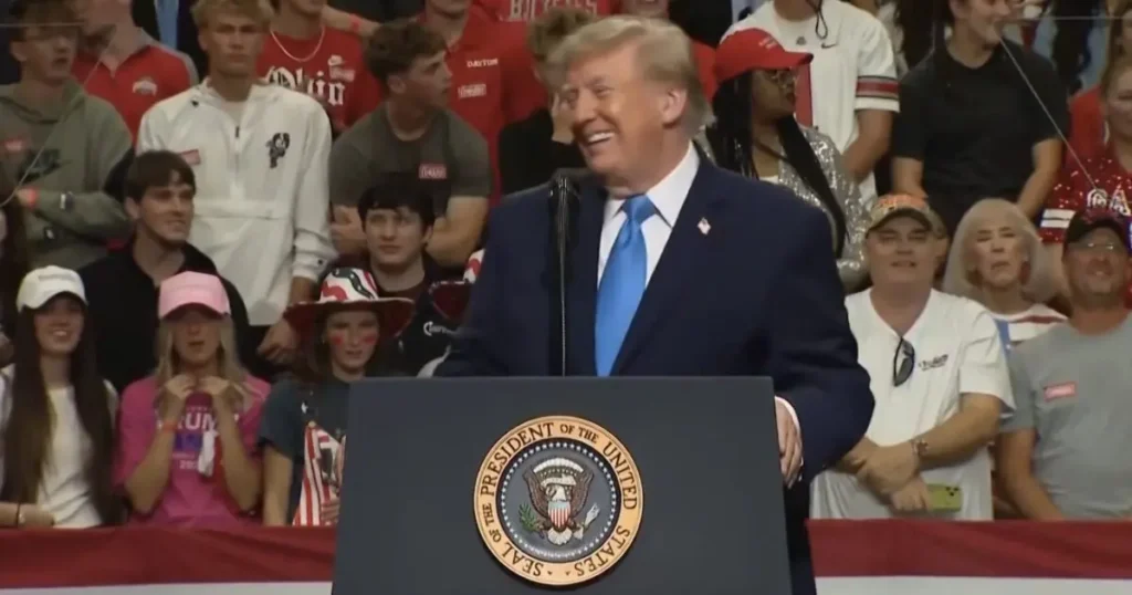 The image shows a political rally with a speaker at a podium featuring the presidential seal, addressing a crowd of supporters.
