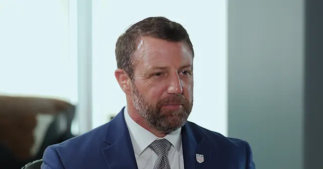 The image shows a man in a professional setting, wearing a blue suit with a white shirt and a patterned tie, and a pin on his lapel.