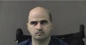 The image appears to be a mugshot of a man seated in a wheelchair, wearing a dark-colored shirt.