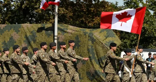 A group of soldiers in camouflage uniforms march in formation, carrying flags, during a military parade.