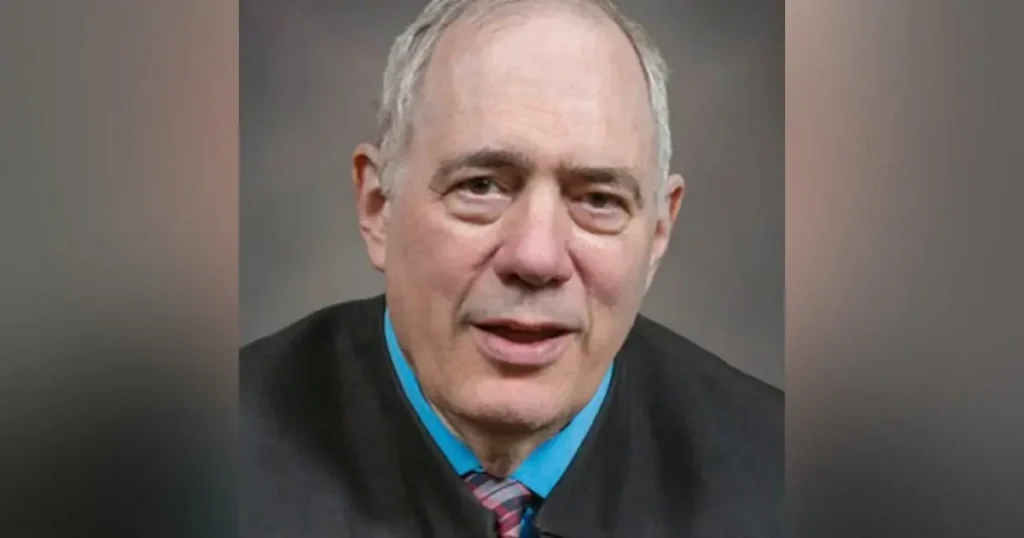 The image shows a man in formal attire, including a suit and tie, speaking in what appears to be a professional or news setting.