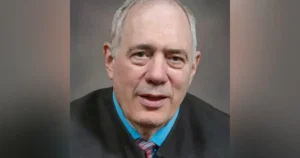 The image shows a man in formal attire, including a suit and tie, speaking in what appears to be a professional or news setting.