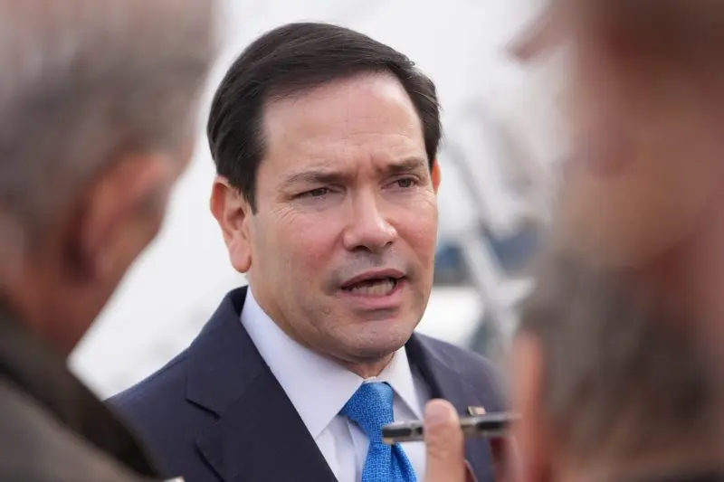The image shows a man in a suit and tie speaking to two other individuals, with a blurred background suggesting an outdoor setting.