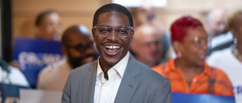 A man in a suit and glasses smiles brightly in front of a group of people at what appears to be a public event or gathering.