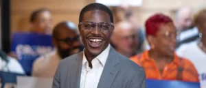 A man in a suit and glasses smiles brightly in front of a group of people at what appears to be a public event or gathering.