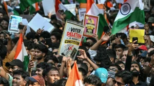 A large crowd of people holds up signs and flags, with one sign reading "Voter Deletion is Against Constitution," indicating a protest or demonstration.