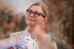 A woman in a professional setting, wearing glasses and a suit, appears to be engaged in a discussion or presentation.