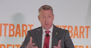 A man in a suit and tie is speaking in front of a backdrop with the word "REITBART" repeated.