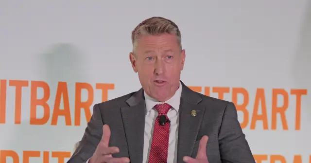 A man in a suit and tie is speaking in front of a backdrop with the word "REITBART" repeated.