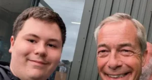 The image shows two individuals standing close together, smiling, with a building and glass window in the background.