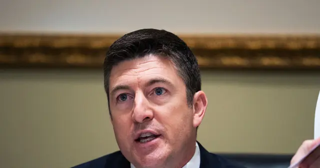 The image shows a man in a suit speaking, with a formal setting in the background.