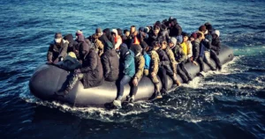 The image shows a group of people on a small inflatable boat in the middle of the ocean, likely indicating a migration or refugee situation.