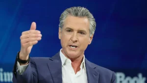 The image shows a man in a suit gesturing with his right hand, likely during a speech or presentation.