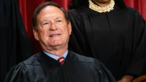 The image shows a person in a judicial robe, seated and smiling, with another individual in a similar robe standing behind them.
