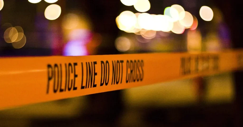The image shows a close-up of a "POLICE LINE DO NOT CROSS" tape at a crime scene with blurred lights in the background.