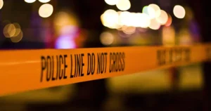 The image shows a close-up of a "POLICE LINE DO NOT CROSS" tape at a crime scene with blurred lights in the background.