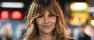 The image shows a person with long, layered hair smiling in a well-lit environment with blurred background lights.