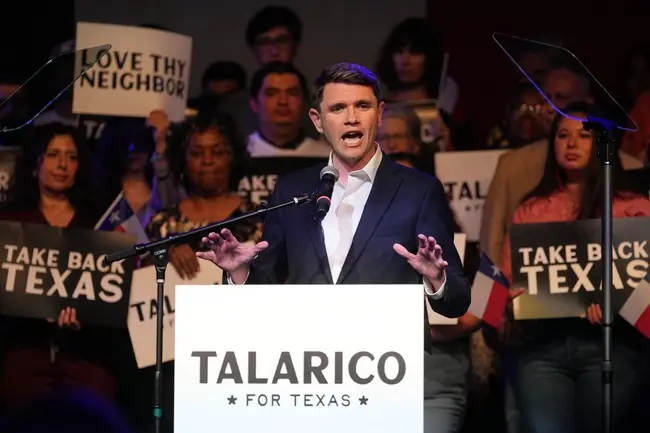 A candidate speaks at a podium with a sign that reads "TALARICO FOR TEXAS" while supporters hold signs in the background.