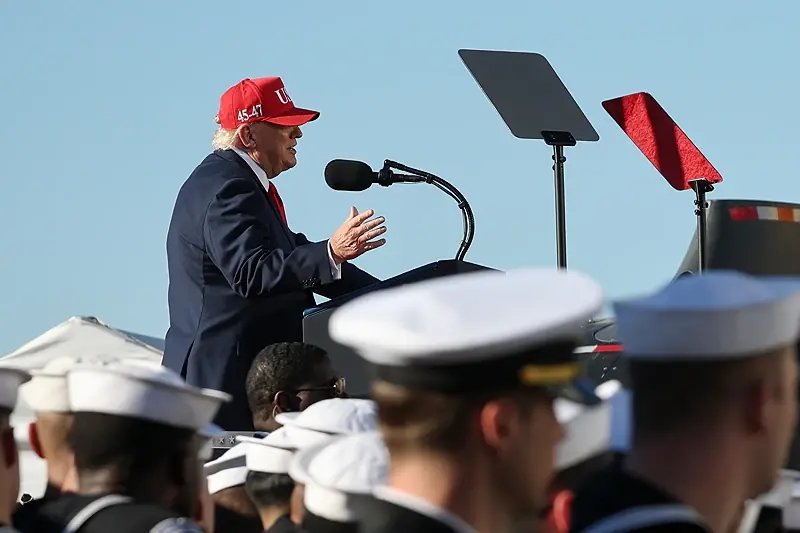 The image shows a person speaking at a podium in front of a group of uniformed individuals, likely during a public event or speech.
