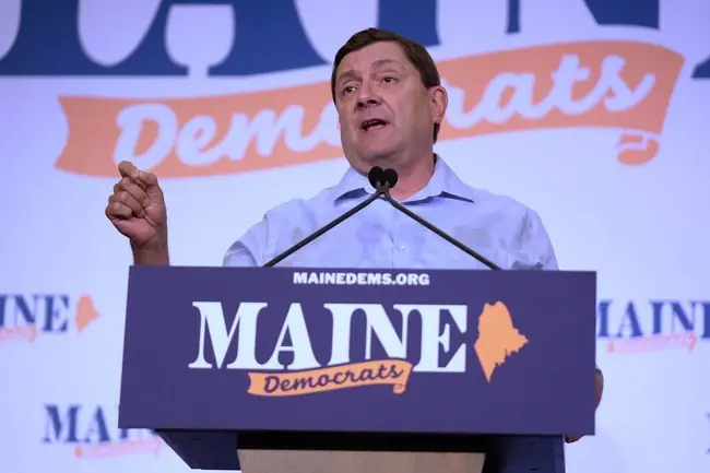 A man is giving a speech at a podium with "Maine Democrats" branding in the background.