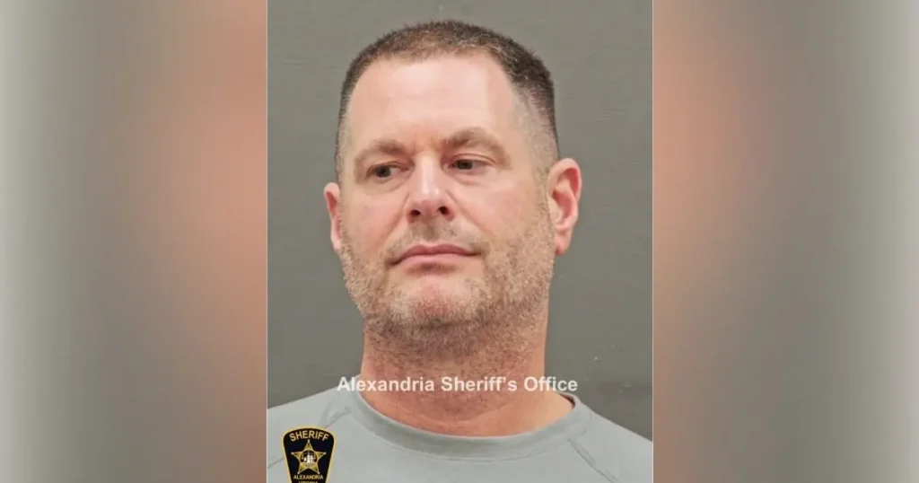 The image is a mugshot from the Alexandria Sheriff's Office, featuring a person in a gray shirt with a sheriff's badge.