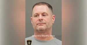The image is a mugshot from the Alexandria Sheriff's Office, featuring a person in a gray shirt with a sheriff's badge.