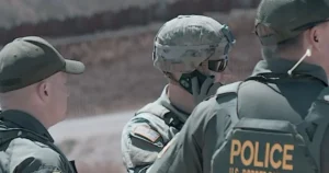 The image shows three individuals in tactical gear, including a person wearing a "POLICE" vest, engaged in a discussion in an outdoor setting.