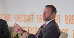 A man in a suit is speaking at a panel discussion with the "Breitbart" logo in the background.