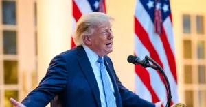 The image shows a person in a suit speaking at a podium with American flags in the background.