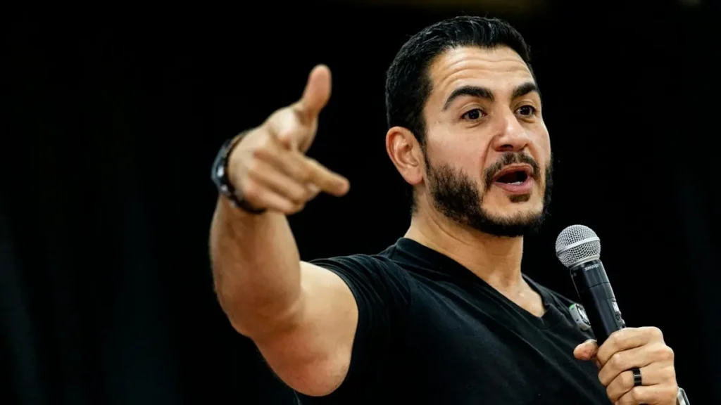 The image shows a man speaking into a microphone while pointing with his other hand, suggesting he is addressing an audience or making a point during a presentation or speech.
