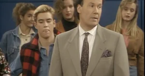 The image shows a man in a suit standing in front of a group of people, possibly in a news or interview setting.