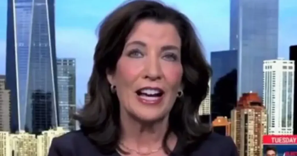 The image shows a news anchor in front of a cityscape backdrop, likely during a broadcast.