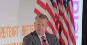The image shows a man in a suit and tie speaking at a podium with an American flag and the word "Breitbart" in the background.