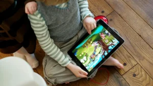 A child is sitting on a wooden floor, holding a tablet displaying a colorful animated game.