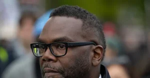 The image shows a close-up of a man wearing glasses, with a blurred background of people and activity.