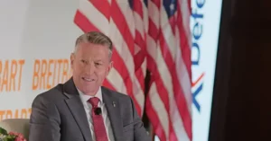 A man in a suit and tie is speaking at a podium with an American flag and a banner in the background.