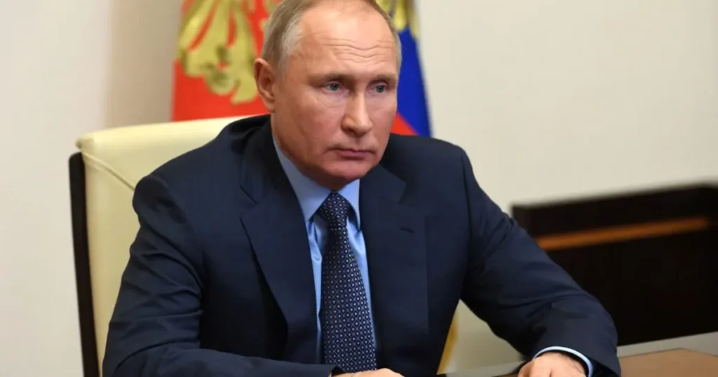 The image shows a person in formal attire seated at a desk, with a flag in the background, suggesting a formal or official setting.