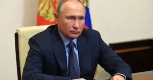 The image shows a person in formal attire seated at a desk, with a flag in the background, suggesting a formal or official setting.