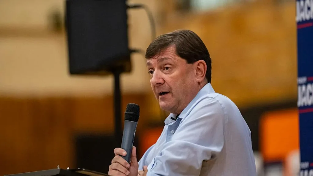 A man is speaking into a microphone at a public event, with campaign signs visible in the background.